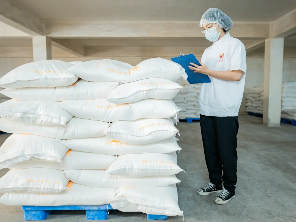 An employee in a lab coat stands at the flour raw material, checking and recording with documents and a pen in hand